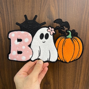 May include: A Halloween-themed patch with the word "BOO" in pink and white, featuring a ghost, pumpkin, spiderweb, and bats. The ghost has a flower on its head. The patch is held against a wooden background.