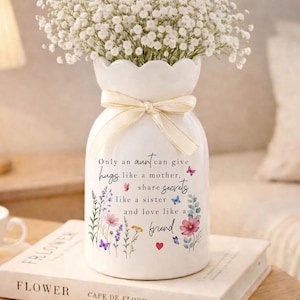 May include: A white ceramic vase with a scalloped rim, tied with a cream-colored ribbon, and filled with white flowers. The vase has a floral and butterfly design with the text: "Only an aunt can give hugs like a mother, share secrets like a sister and love like a friend."