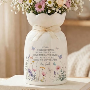 Puede incluir: Un jarrón de cerámica blanco con un arreglo floral. El jarrón tiene una cinta color crema y presenta un diseño floral y de mariposas con el texto: "NUNCA SUBESTIMES LA DIFERENCIA QUE HAS HECHO Y LAS VIDAS QUE HAS TOCADO. DISFRUTA DEL PRÓXIMO CAPÍTULO Sra. Smith."