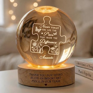 Op de afbeelding: Een verlichte, bolvormige glazen ornament met een houten basis. De glazen bol heeft een puzzelstukontwerp met de tekst "Never Forget You Are The Piece That Made The Difference" en de naam "Amanda". De houten basis leest "Thank You For Being Such An Important Piece In Our Team".