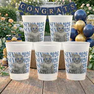 May include: Five white plastic cups with a photo of a child wearing heart-shaped sunglasses, the text "SENIOR 2026" and the name "Sophia" in blue. A banner that says "CONGRATS" hangs in the background.