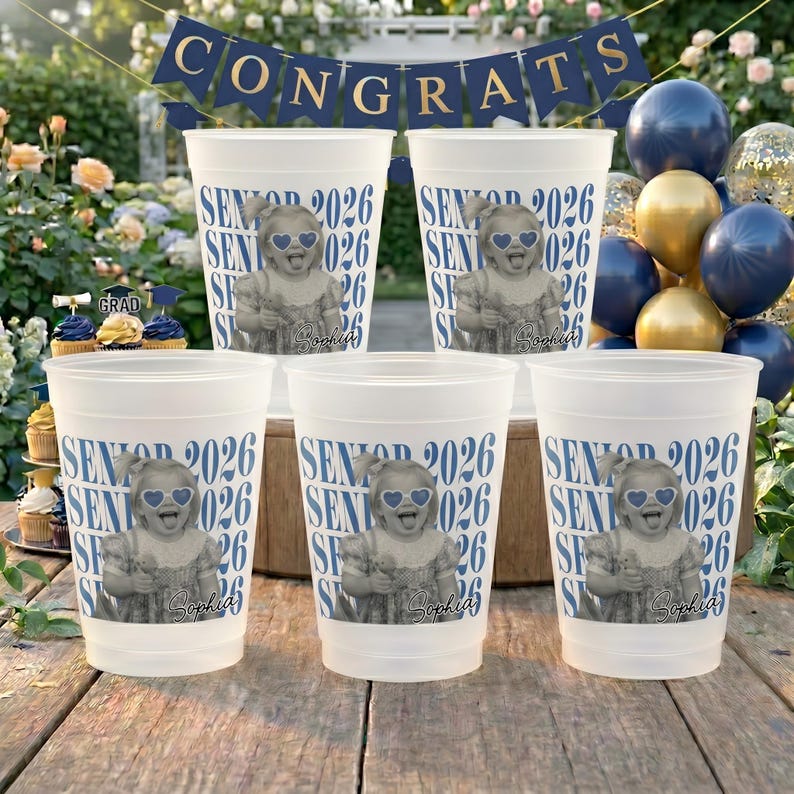 K&ouml;nnte beinhalten: F&uuml;nf wei&szlig;e Plastikbecher mit einem Abschluss-Thema. Jeder Becher zeigt ein Foto eines Kindes mit herzf&ouml;rmiger Sonnenbrille, den Text "SENIOR 2026" und den Namen "Sophia". Ein Banner mit der Aufschrift "CONGRATS" h&auml;ngt im Hintergrund.