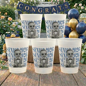 Puede incluir: Seis vasos de plástico blanco. Cada vaso presenta una imagen en blanco y negro de un bebé con gafas de sol y pajarita, con el texto "SENIOR 2026" en azul y el nombre "James".