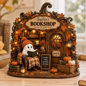 Peut inclure: Une décoration de table représentant un fantôme avec un bonnet orange, assis dans un fauteuil lisant un livre dans une structure en forme de citrouille marquée Sophia's BOOKSHOP EST. 2009. Elle inclut des citrouilles, des livres et des lumières.