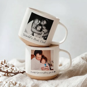 Pode incluir: Duas canecas de cerâmica branca empilhadas. Cada caneca tem uma foto impressa. A caneca superior diz "World's Best Dad" e a inferior "Grateful for everything you do". A base de cada caneca é castanha.