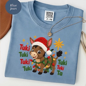 May include: A light blue t-shirt with an embroidered donkey wearing a Santa hat and Christmas lights. The text "Tuki Tuki" is embroidered in red and green. A gold necklace is draped across the shirt.