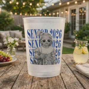 K&ouml;nnte beinhalten: Ein wei&szlig;er Plastikbecher mit blauem Aufdruck "Senior 2026" und einem Schwarz-Wei&szlig;-Foto eines jungen M&auml;dchens mit herzf&ouml;rmiger Sonnenbrille. Der Name "Sophia" ist in Schreibschrift geschrieben.