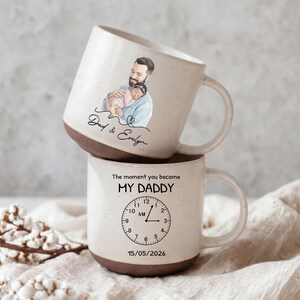 Op de afbeelding: Twee gestapelde keramische mokken met een gespikkelde beige afwerking en bruine basis. De bovenste mok heeft een aquarel illustratie van een vader die een baby vasthoudt, met de tekst "Dad & Evelyn". De onderste mok heeft een klokafbeelding en de tekst "The moment you became MY DADDY 15/05/2026."