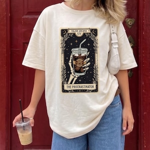 May include: An off-white short-sleeve t-shirt featuring a graphic of a skeleton hand holding an iced coffee in a tarot card style. The card is labelled Major Arcana XIV and The Procrastinator, set against a dark background with stars.