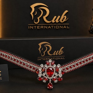 May include: A black leather browband featuring a central ornate cluster of red and clear crystals, flanked by two rows of red crystals set in silver-toned metal. The piece is displayed in front of a black box with the gold text Rub International.