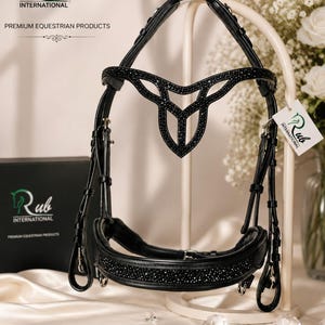 May include: A black leather horse bridle featuring a browband with a decorative, symmetrical cutout design encrusted with small black crystals. The noseband is also embellished with a row of black crystals. A white tag displays the Rub International logo.