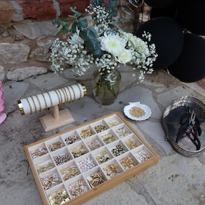 May include: A display of jewelry-making supplies, including a wooden tray with various charms, a bracelet display, and a pink sign. A vase of flowers and black balloons are in the background. The charms are gold, silver, and pearl.