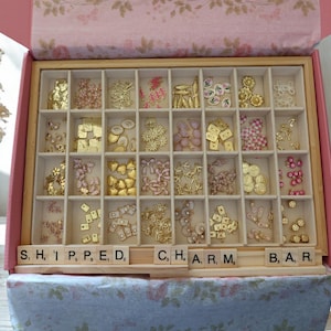 May include: A wooden organizer box with twenty-eight compartments filled with assorted small metal and resin charms in gold, pink, and white tones. The box features the text S, H, I, P, P, E, D, C, H, A, R, M, B, A, R, on small wooden letter tiles.