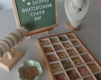 May include: A charm bar display featuring a green felt letter board with white text, a necklace on a mannequin bust, a wooden bracelet stand with gold chains, a wooden organizer tray with various charms, a small bowl of jump rings, and pink pliers.