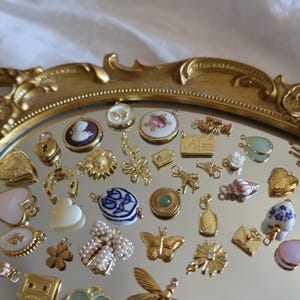 May include: A collection of small, ornate charms and pendants in gold tones, including hearts, butterflies, and floral designs, arranged on a mirrored surface within a decorative gold-colored tray.