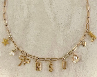 May include: A gold chain necklace with charms, including a star, a pearl, a bow, the letters M, S, and U, a seashell, a heart, and a boot.