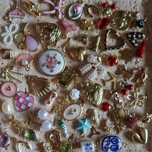 May include: A collection of small, decorative charms in gold, pink, white, and red. The charms include hearts, flowers, bows, and other shapes. Some charms have pearl accents. The charms are likely for jewelry making or crafting.