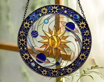 May include: A circular stained glass suncatcher featuring a central yellow sun surrounded by orbiting planets and rings. The outer border is a deep blue frame decorated with yellow stars and small dark spheres, suspended by a black metal chain.