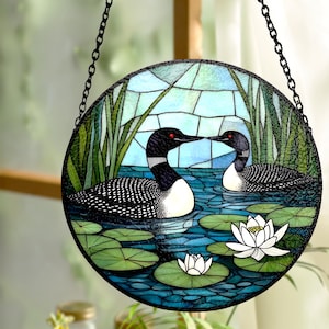 May include: Round stained glass panel featuring two loons on a blue pond with lily pads and white flowers. The loons have black and white plumage, red eyes, and are set against a backdrop of green reeds and a light blue sky. The panel is suspended by a black chain.