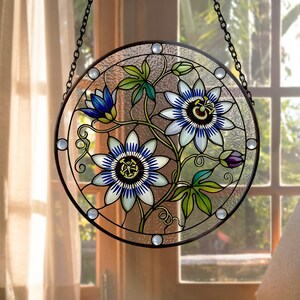 May include: A circular stained glass suncatcher featuring two large passion flowers with white and blue petals, a smaller blue bud, and green leaves. The design is enclosed in a dark metal frame with small clear glass accents and a hanging chain.