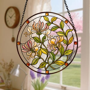 May include: A circular stained glass suncatcher featuring a honeysuckle floral design with pink, yellow, and white petals and green leaves. The piece is framed in a dark metal border with small amber glass accents and hangs from a black metal chain.