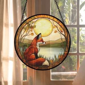 Peut inclure: Vitrail rond représentant un renard assis au bord d'un lac sous une pleine lune. Le renard a un pelage orange et blanc, et la scène comprend des arbres et des feuilles d'automne. L'œuvre est encadrée en noir et suspendue à une chaîne.