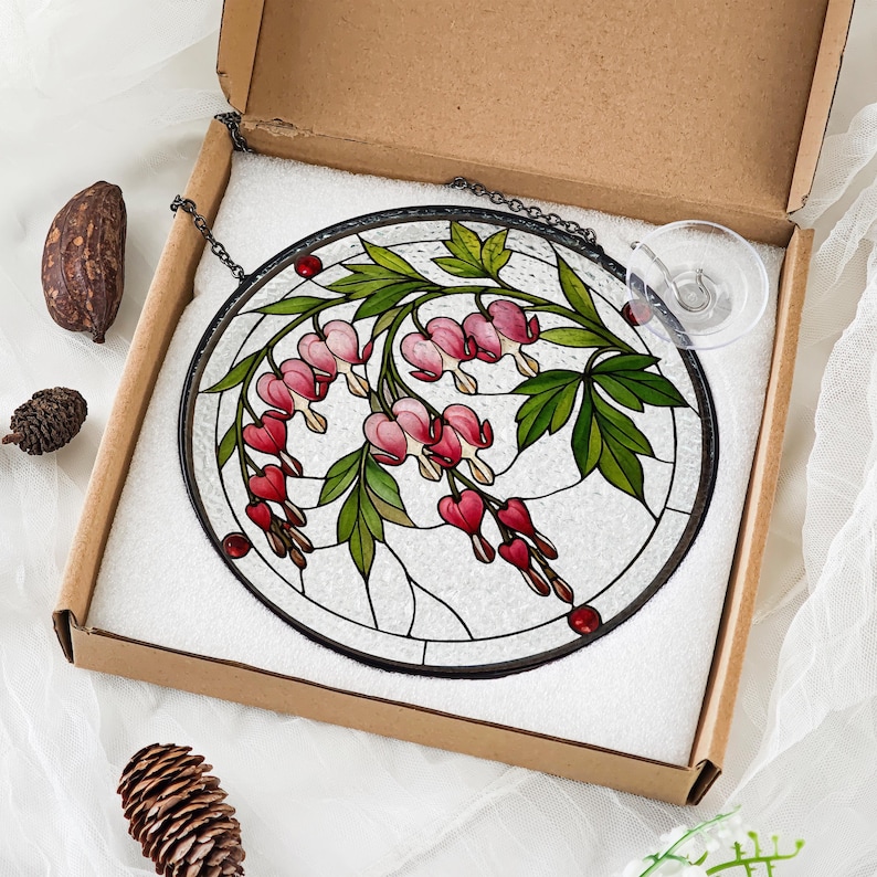 May include: A circular stained glass suncatcher featuring a botanical design of pink bleeding heart flowers and green leaves against a textured clear glass background, framed in black metal with a hanging chain and a clear suction cup attachment.