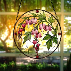 Op de afbeelding: Een ronde glas-in-lood zonnevanger met een botanisch ontwerp van roze gebroken hartjesbloemen met groene bladeren. Het stuk is gevat in een donkere metalen rand met kleine rode glazen accenten en hangt aan een zwarte metalen ketting.