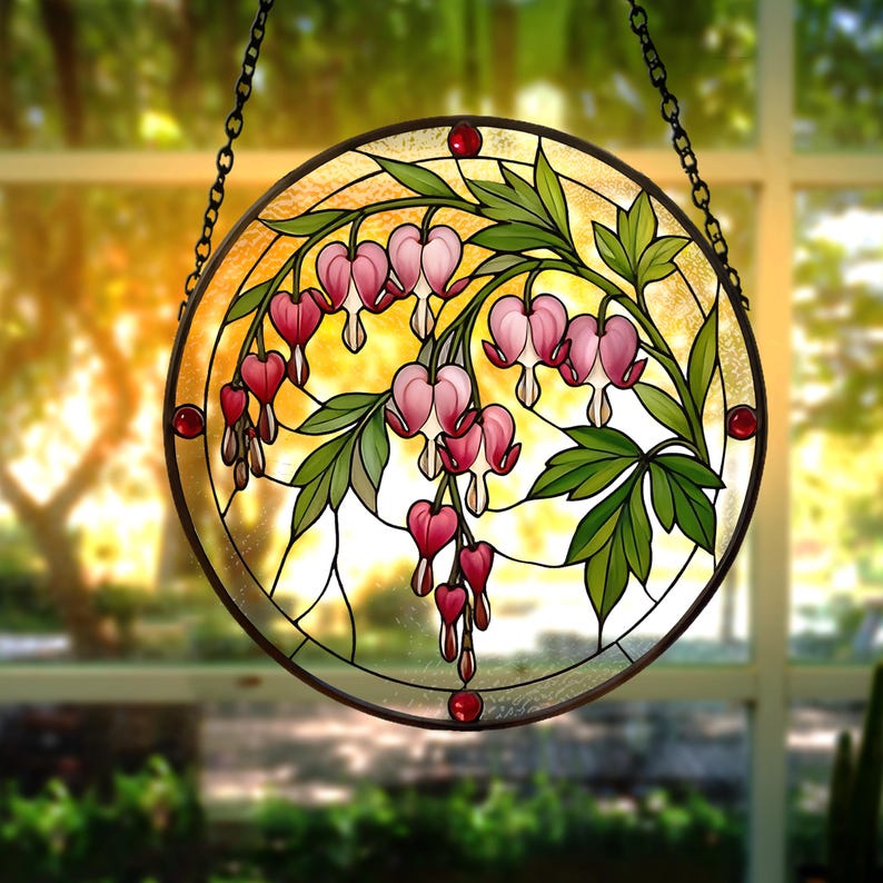 May include: A circular stained glass suncatcher featuring a botanical design of pink bleeding heart flowers with green leaves. The piece is framed in a dark metal border with small red glass accents and is suspended by a black metal chain.