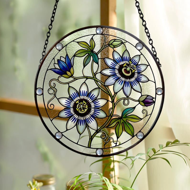 May include: A circular stained glass suncatcher featuring a detailed illustration of blue and white passion flowers with green leaves. The piece is framed in a dark metal border with small circular accents and hangs from a black metal chain.