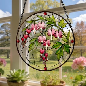 May include: A circular stained glass suncatcher featuring a botanical design of pink and red bleeding heart flowers with green leaves. The piece is framed in a dark metal border and suspended by a black chain, showcasing intricate glass segments.