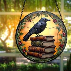 May include: Round stained glass window art featuring a black crow perched atop a stack of books. The window has a yellow and orange background with green vines and red berries around the edge. A black chain is attached for hanging.