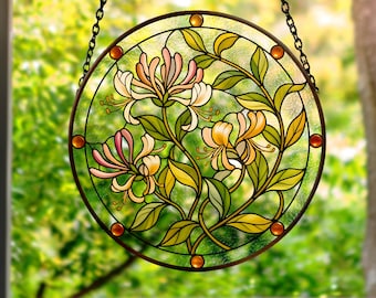May include: A circular stained glass suncatcher featuring a honeysuckle floral design with pink, yellow, and green glass segments. The piece is framed in a dark metal border with small amber-colored glass accents and hangs from a black metal chain.