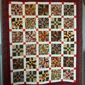May include: A patchwork quilt with a burgundy border and orange squares. The quilt is made up of various autumnal fabrics featuring pumpkins, sunflowers, and leaves.