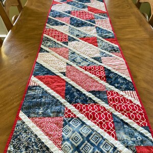 May include: A red, white, and blue quilted table runner with a diagonal pattern. The runner is made of various fabrics with different patterns, including stripes, polka dots, and geometric designs.