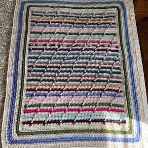 May include: A handmade crochet blanket with a geometric pattern in various colors, including blue, pink, brown, and green. The blanket has a cream-colored border with blue and green stripes. The overall design is a mix of textures and colors.