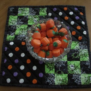 May include: A black and green quilted table runner with a white spiderweb design. A clear glass bowl filled with orange candy corn sits on the runner.