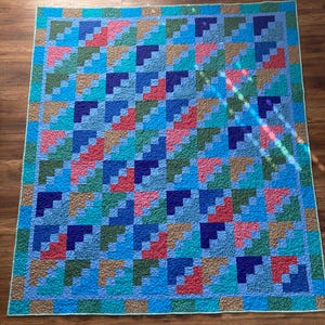 May include: A handmade quilt with a geometric pattern in shades of blue, green, pink, and brown. The quilt features a repeating block design, creating a visually appealing and colorful textile. The edges are finished with a light green border.