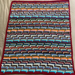 May include: A handmade, colorful crochet blanket with a repeating pattern of horizontal stripes. The blanket features a wide variety of colors, including blue, orange, green, and brown. The border is a solid burgundy color.