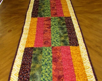 May include: A quilted table runner with a patchwork design featuring various shades of brown, green, red, and yellow. The runner has a white and gold border with a decorative pattern.
