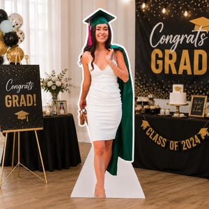 May include: A life-size cardboard cutout of a graduate wearing a white sleeveless dress, a green graduation cap with a pink tassel, and a green graduation gown draped over one shoulder. The graduate is holding a pair of shoes in one hand.