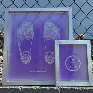 May include: Two purple screen printing frames with white designs. The larger frame has a design of two bare footprints. The smaller frame has a design of a person wearing a hat and a coat with the text "2019" and "10x12".