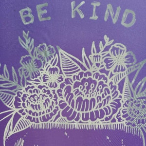 May include: Purple background with a white skull design featuring a floral crown. The text "BE KIND" is written above the skull.