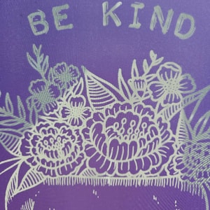 May include: Purple background with a white skull design with a floral crown. The text "BE KIND" is written above the skull.