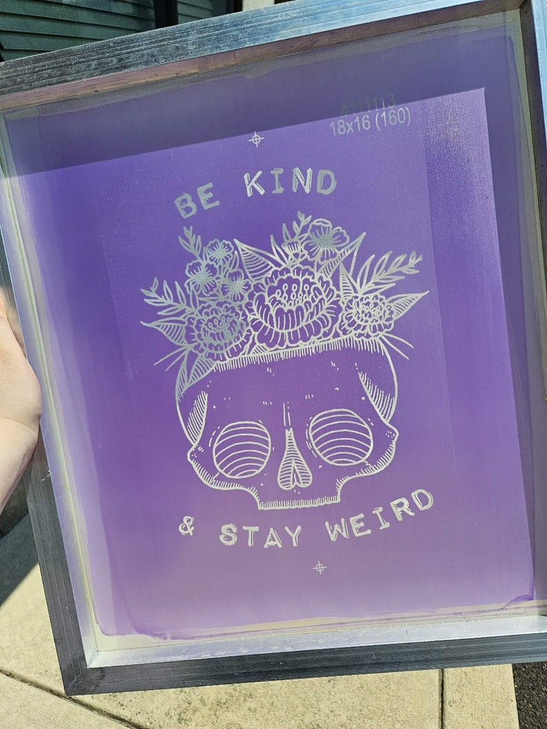 May include: A purple screen printing frame with a white design that says "Be Kind & Stay Weird" with a skull and floral design.