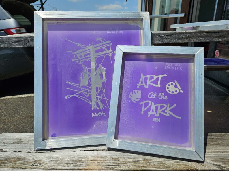May include: Two purple screen printing frames with silver frames. The larger frame has the text "ART at the PARK 2024" with a paint palette and a leaf design. The smaller frame has a drawing of a utility pole with wires and the text "kimfitz".