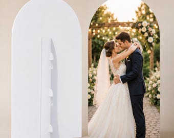 May include: A white arched display board featuring a vertical support stand on the back. The front surface displays a high-resolution photograph of a couple in wedding attire embracing on a stone path surrounded by floral arrangements and lights.