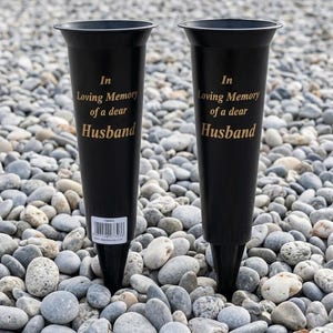 May include: Two black memorial vases with a flared top and pointed base. Gold text reads "In Loving Memory of a dear Husband". The vases are displayed on a bed of grey and white pebbles.