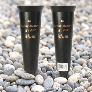 Puede incluir: Dos jarrones conmemorativos negros con la parte superior acampanada, inscritos con letras doradas que dicen "In Loving Memory of a dear Mum". Los jarrones están colocados sobre un lecho de guijarros lisos grises y blancos.