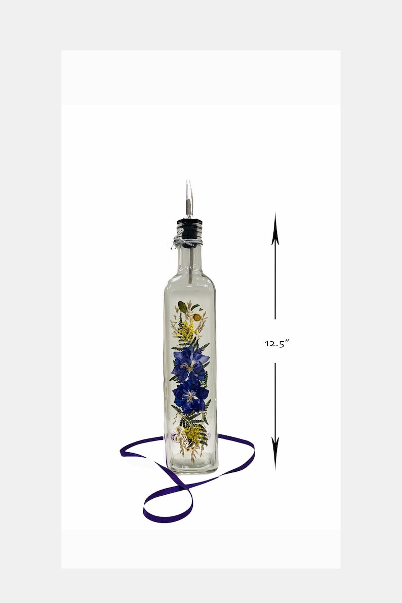 Puede incluir: Una botella de vidrio transparente con un pico de vertido de metal y un dise&ntilde;o floral con flores prensadas azules, amarillas y verdes. La botella mide 12,5 pulgadas de alto.